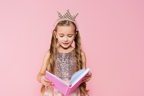 happy little girl in crown reading book isolated on pink
