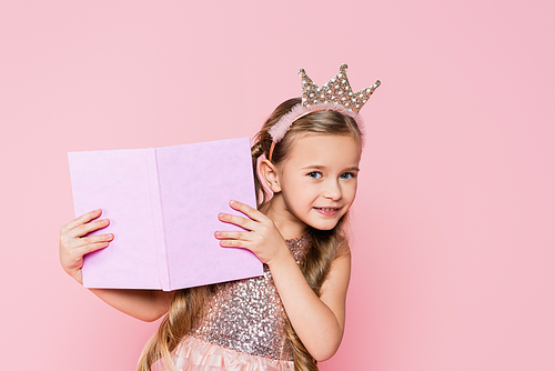 happy little girl in crown holding book isolated on pink