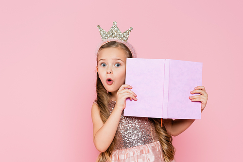 surprised little girl in crown holding book isolated on pink