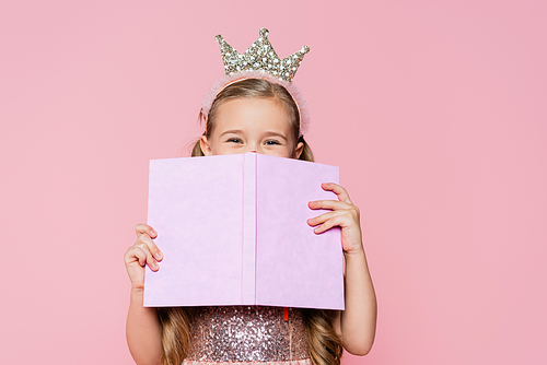 little girl in crown covering face with book isolated on pink