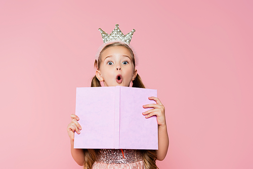 shocked little girl in crown holding book while  isolated on pink