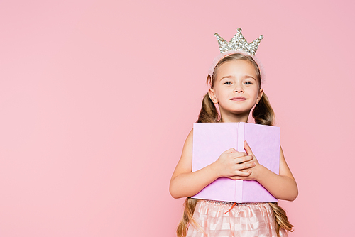 smiling little girl in crown holding book while  isolated on pink