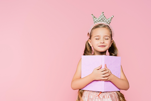 smiling little girl with closed eyes in crown holding book isolated on pink