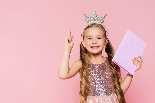 smiling little girl in crown holding book and pointing with finger isolated on pink