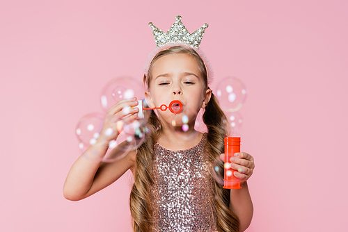 little girl in crown blowing soap bubbles on blurred foreground isolated on pink