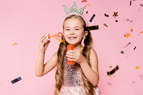 happy little girl in crown holding soap bubbles near falling confetti on pink