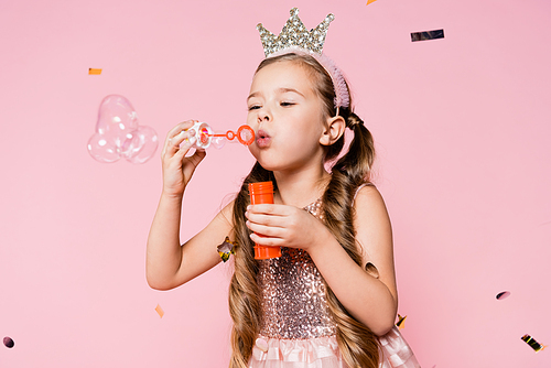 little girl in crown blowing soap bubbles near falling confetti on pink