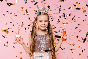 cheerful little girl in crown holding soap bubbles near falling confetti on pink
