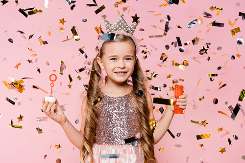 cheerful little girl in crown holding soap bubbles near falling confetti on pink
