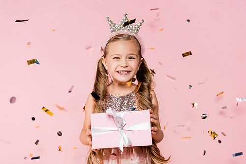 happy little girl in crown holding present near falling confetti on pink