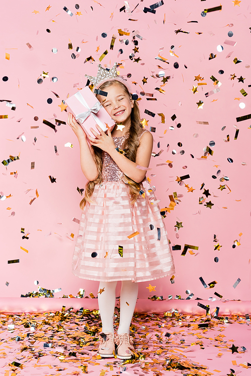 full length of happy little girl in crown holding present near falling confetti on pink