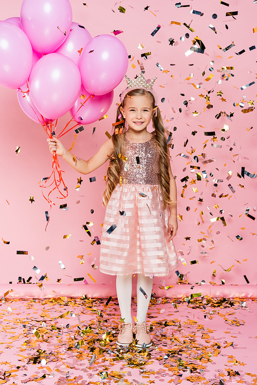full length of happy little girl in crown holding balloons near falling confetti on pink