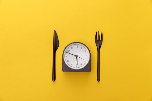 top view of clock between knife and fork on yellow background