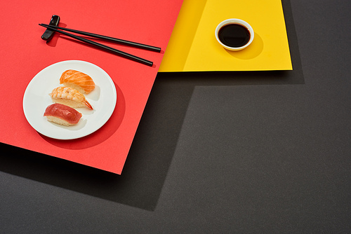 fresh nigiri with salmon, shrimp and tuna near soy sauce and chopsticks on red, yellow and black surface