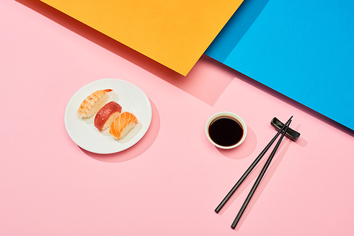 fresh nigiri with salmon, tuna and shrimp near soy sauce and chopsticks on blue, pink, orange surface