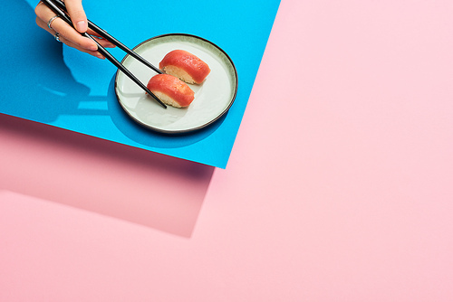 partial view of woman eating fresh nigiri with tuna with chopsticks on blue, pink background