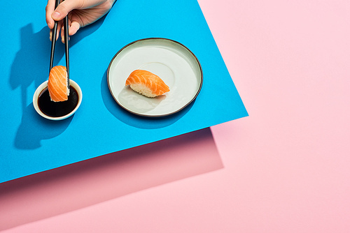 cropped view of woman putting fresh nigiri with salmon into soy sauce on blue, pink background