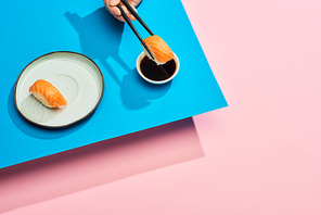 cropped view of woman putting fresh nigiri with salmon into soy sauce on blue, pink background