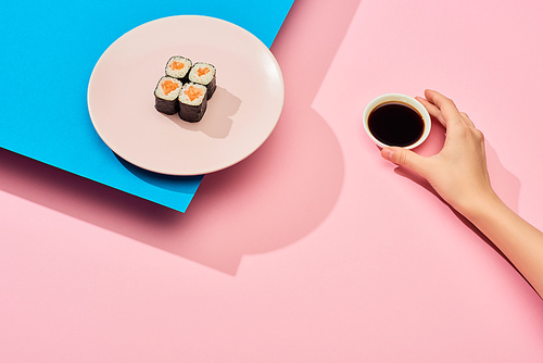 cropped view of woman holding soy sauce near fresh maki with salmon on blue, pink background