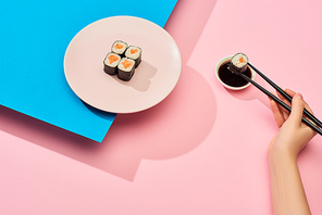 cropped view of woman eating fresh maki with salmon near soy sauce on blue, pink background