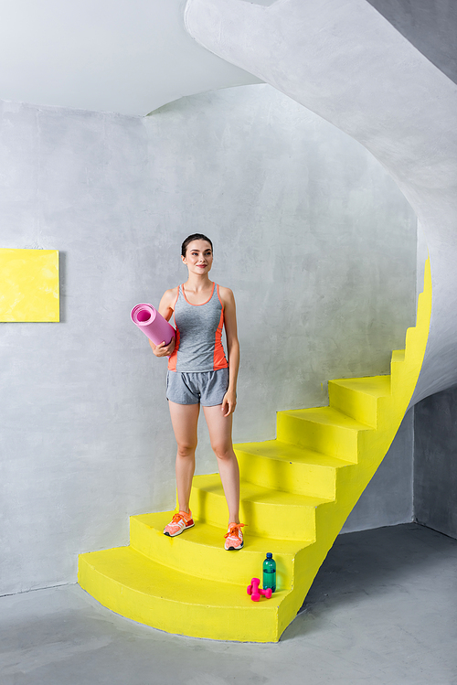 Young woman in sportswear holding fitness mat near bottle of water and dumbbells on stairs at home