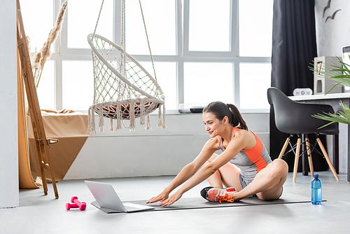 Selective focus of young woman warming up near laptop on fitness mat at home