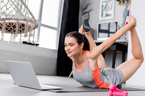 Selective focus of young sportswoman exercising on fitness mat near laptop and dumbbells at home