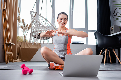 Young woman looking at laptop while training on fitness mat near dumbbells at home