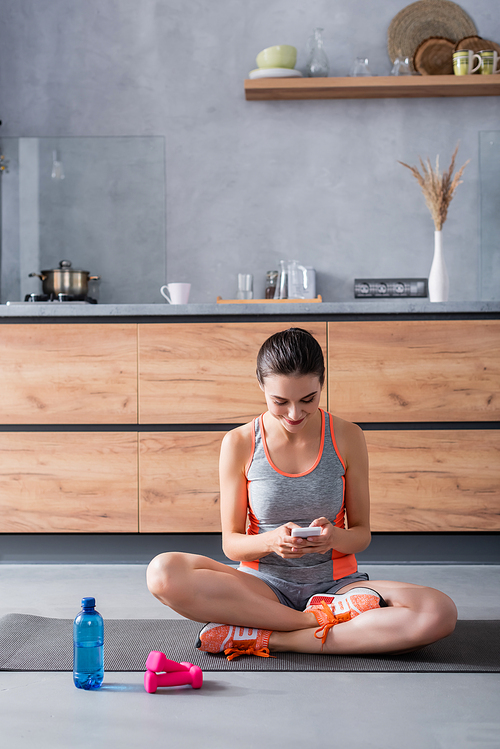 Sportswoman using smartphone on fitness mat near dumbbells and bottle of water at home