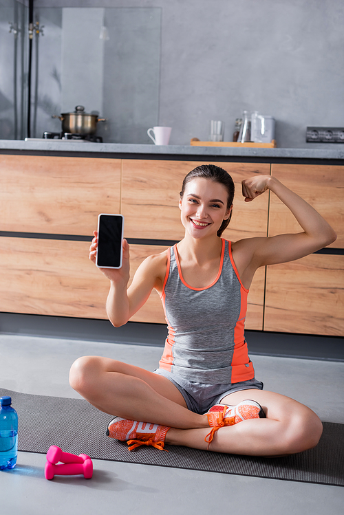 Sportswoman showing smartphone with blank screen near bottle of water and sport equipment in kitchen