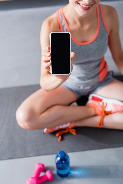 Cropped view of woman in sportswear and sneakers showing smartphone with blank screen near bottle of water and sport equipment