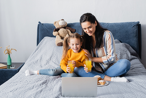 cheerful woman with daughter holding orange juice while watching film on laptop
