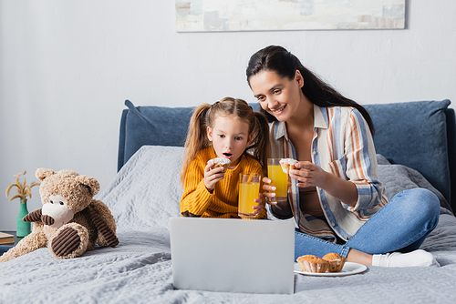 excited child with happy mother watching movie on laptop with orange juice and muffins