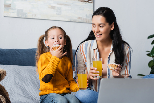 daughter eating delicious muffin while watching film on computer with mother