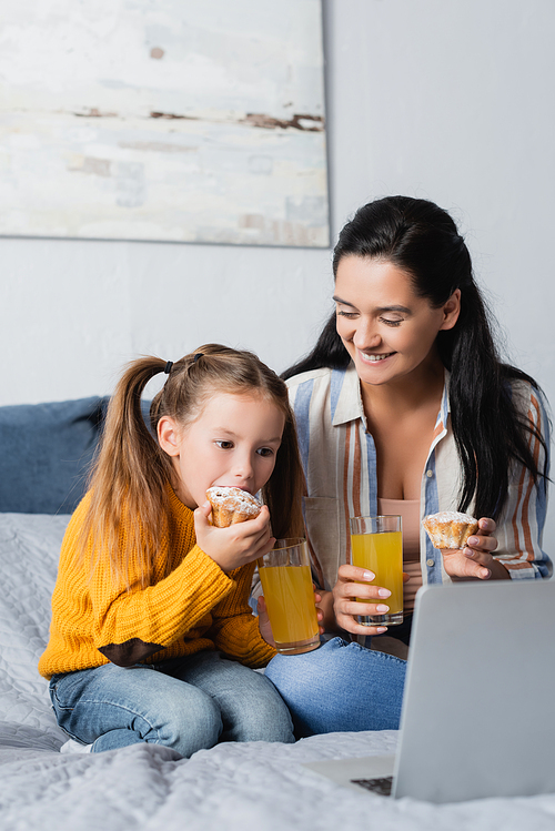happy mother looking at daughter eating muffin and watching movie on laptop