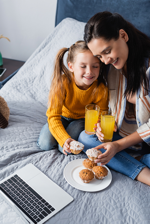 happy mother and daughter holding muffin and orange juice near laptop
