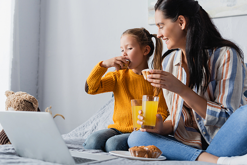 happy mother looking at daughter eating muffin while watching film on laptop