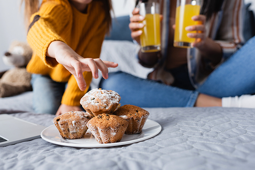 cropped view of child taking muffin from plate near mom holding orange juice on blurred background