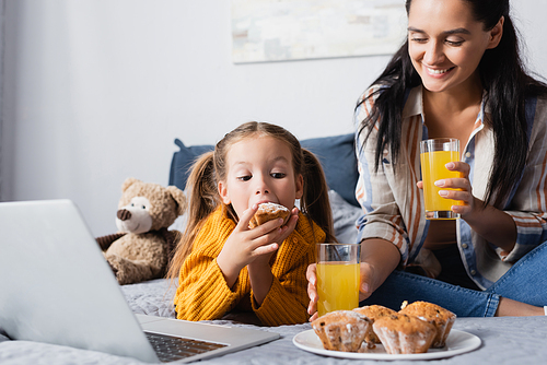 girl eating muffin near cheerful mother while watching movie on laptop