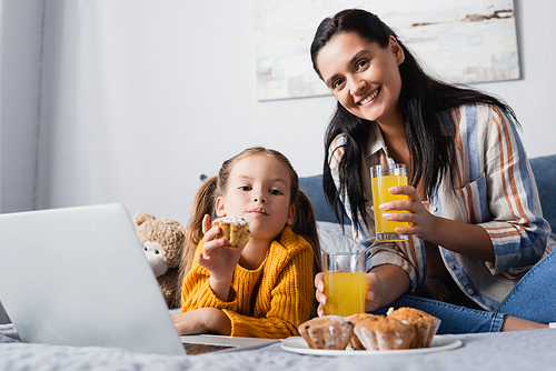 happy woman and daughter  near muffins and orange juice while watching film on laptop