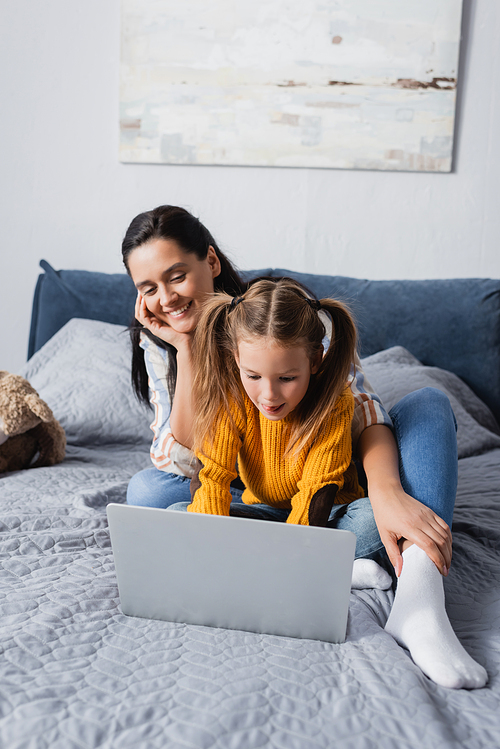 happy mother looking at daughter using laptop in bedroom