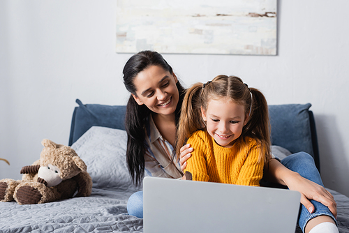 cheerful woman looking at daughter using laptop in bedroom