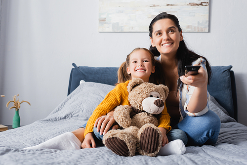 joyful woman holding tv remote controller while watching movie with daughter