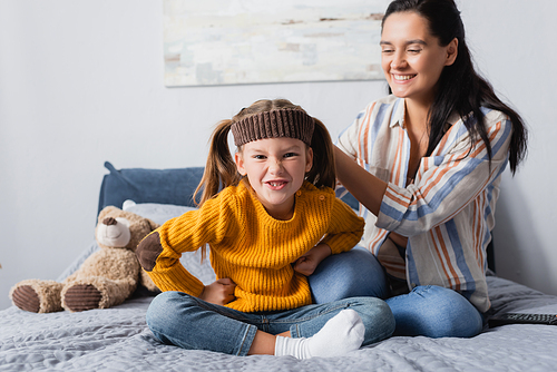 grimacing girl in headband  while sitting near mother on bed