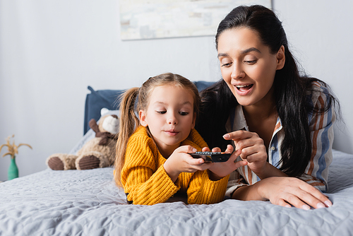 happy mother showing daughter how to use tv remote controller while lying on bed