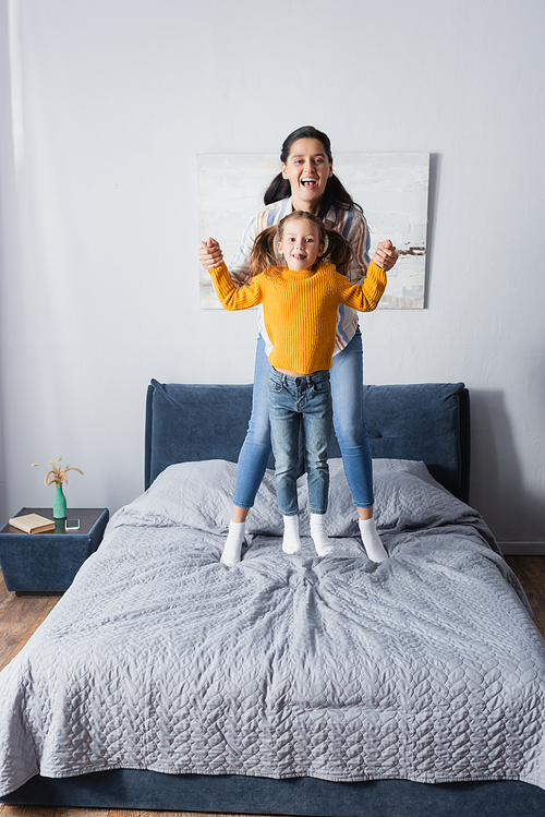 cheerful mother and daughter holding hands while jumping on bed