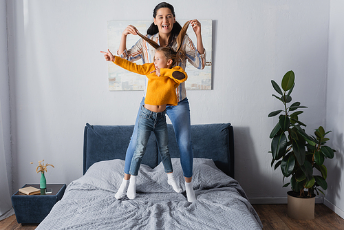 excited mother holding hair of daughter while she pointing with finger and jumping on bed