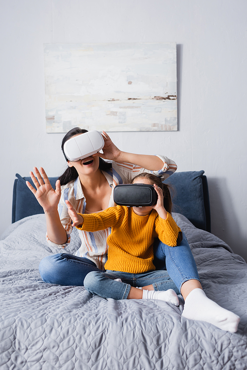mother and child using vr headsets while sitting on bed and gesturing