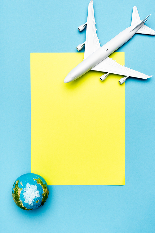 top view of white plane model, globe and empty yellow paper on blue background
