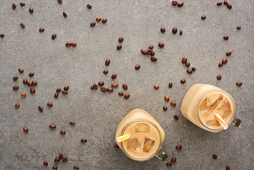 top view of ice coffee in glass jars with straws and scattered coffee grains on grey background